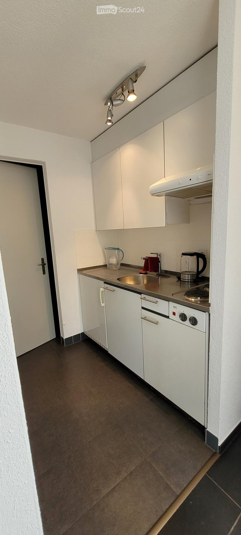 Compact kitchen with white cabinets, stainless steel sink, stove, and oven. Includes a cup, kettle, and towel on the counter.