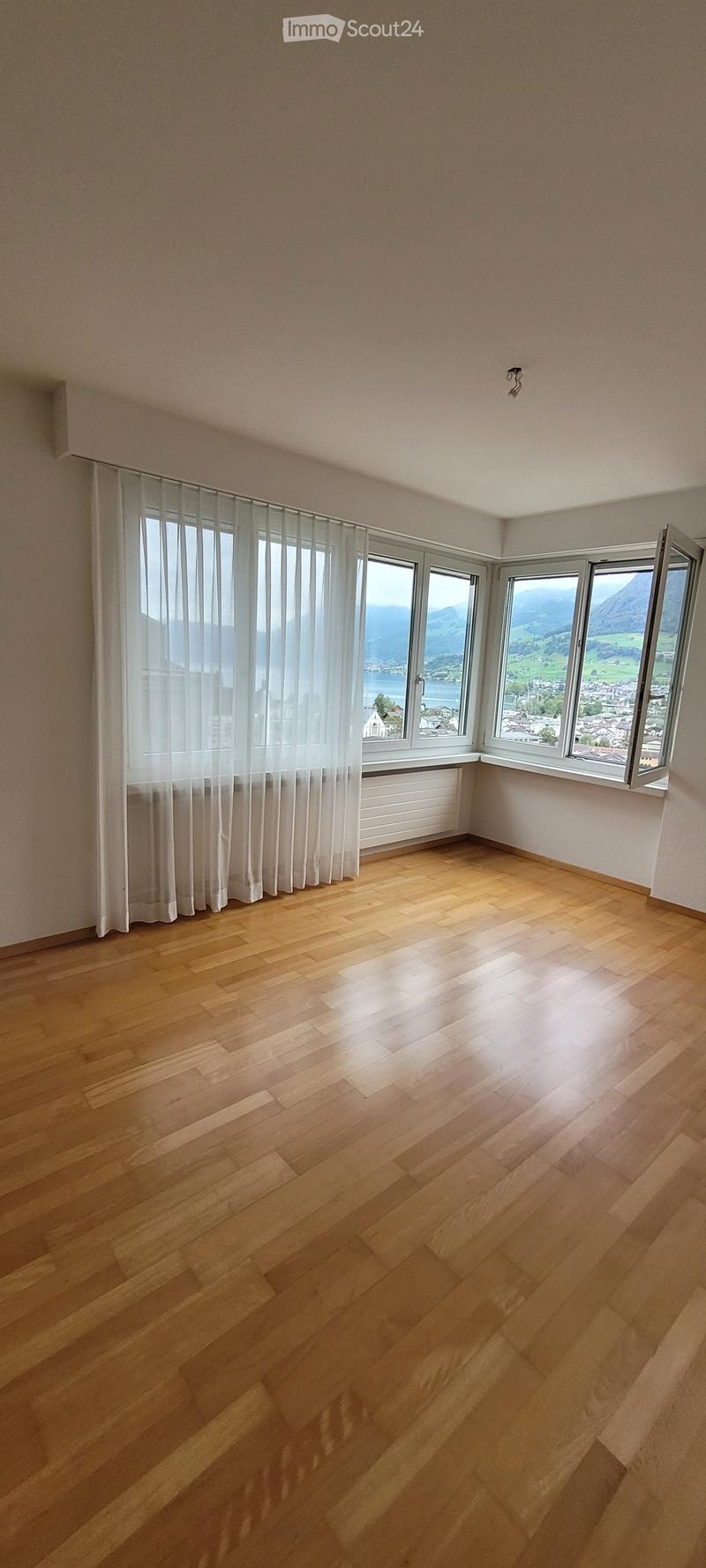Empty room with wooden flooring, large windows with white curtains, and a scenic mountain view.