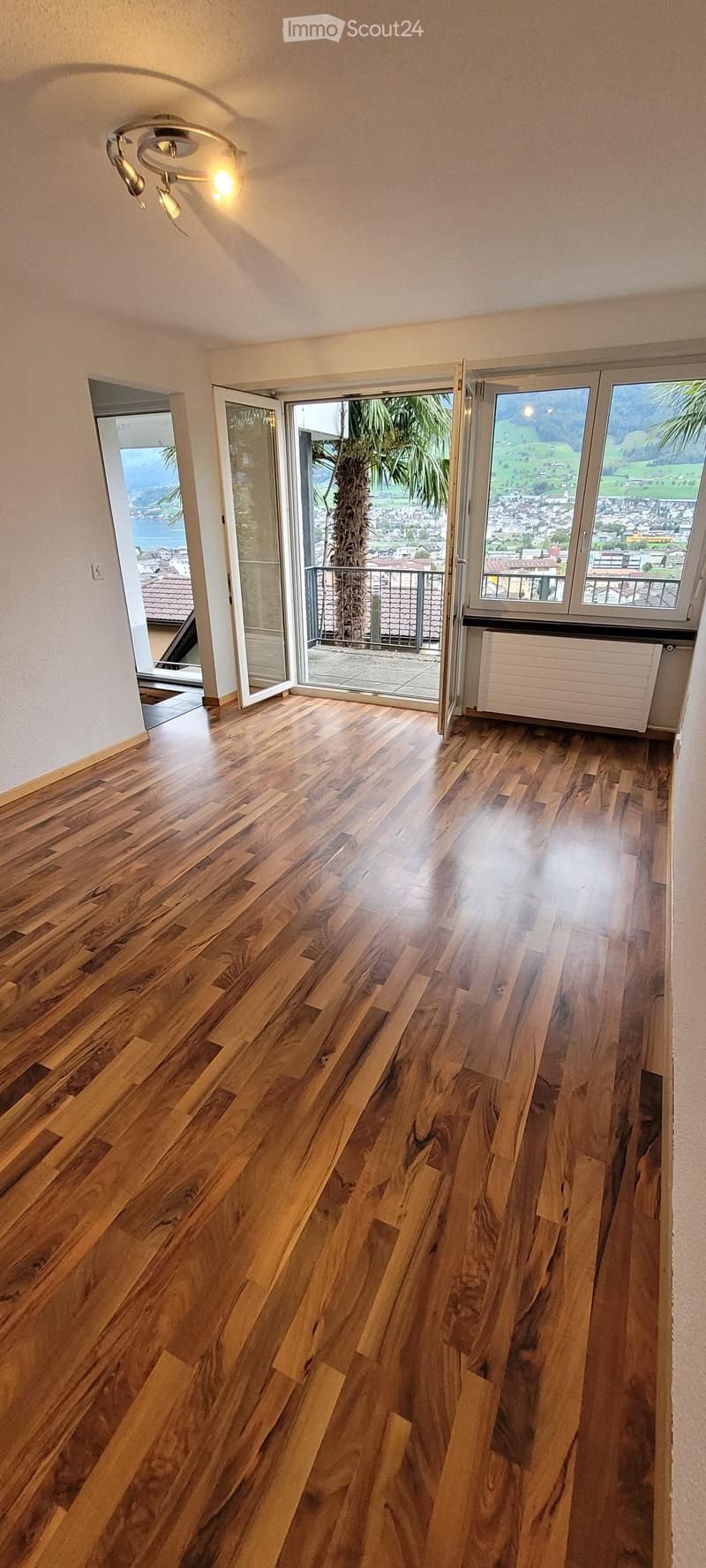 Empty room with polished wooden flooring, large windows, and sliding glass doors leading to a balcony with scenic views.