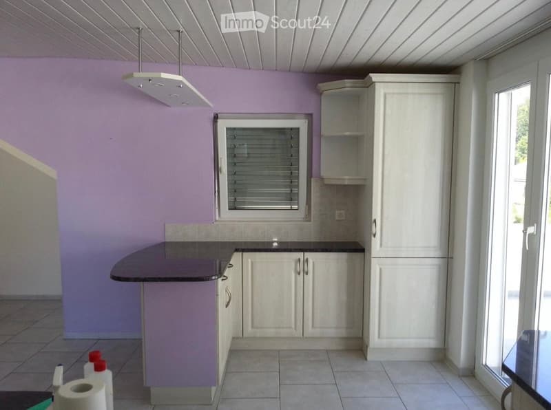 Light purple kitchen with white cabinets, a dark countertop, and a window with blinds. A light fixture hangs from the ceiling.