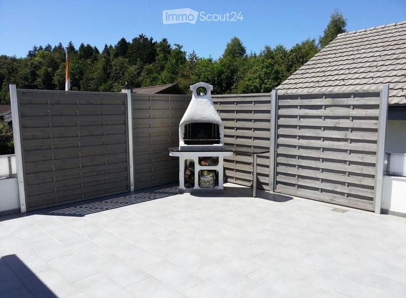 A well-maintained outdoor patio with a wooden fence, a white barbecue, and a small wooden table.