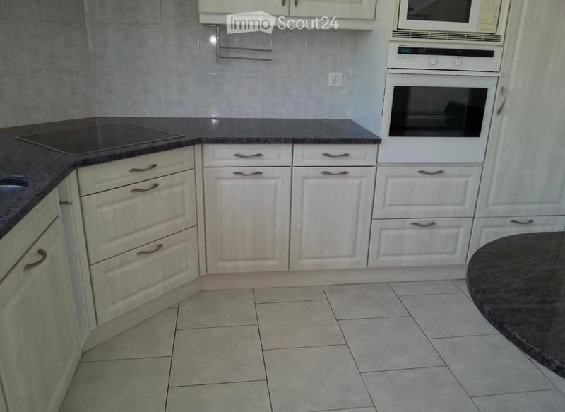 Modern kitchen with white cabinets, a microwave, an oven, and a countertop. The floor is tiled, and a round table is visible in the corner.