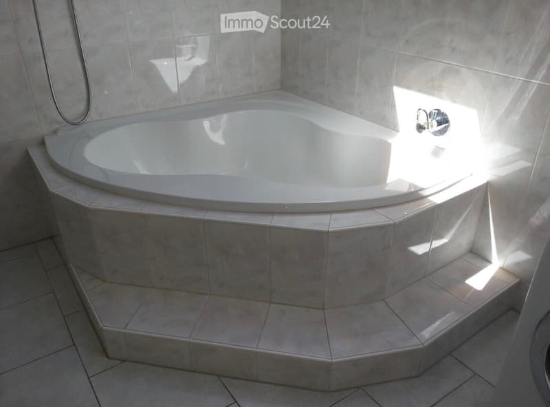 Modern bathroom with a corner bathtub, tiled walls, and a tiled floor. The bathtub has a smooth, curved design and is surrounded by a tiled platform. Natural light streams in from a window, illuminating the space.