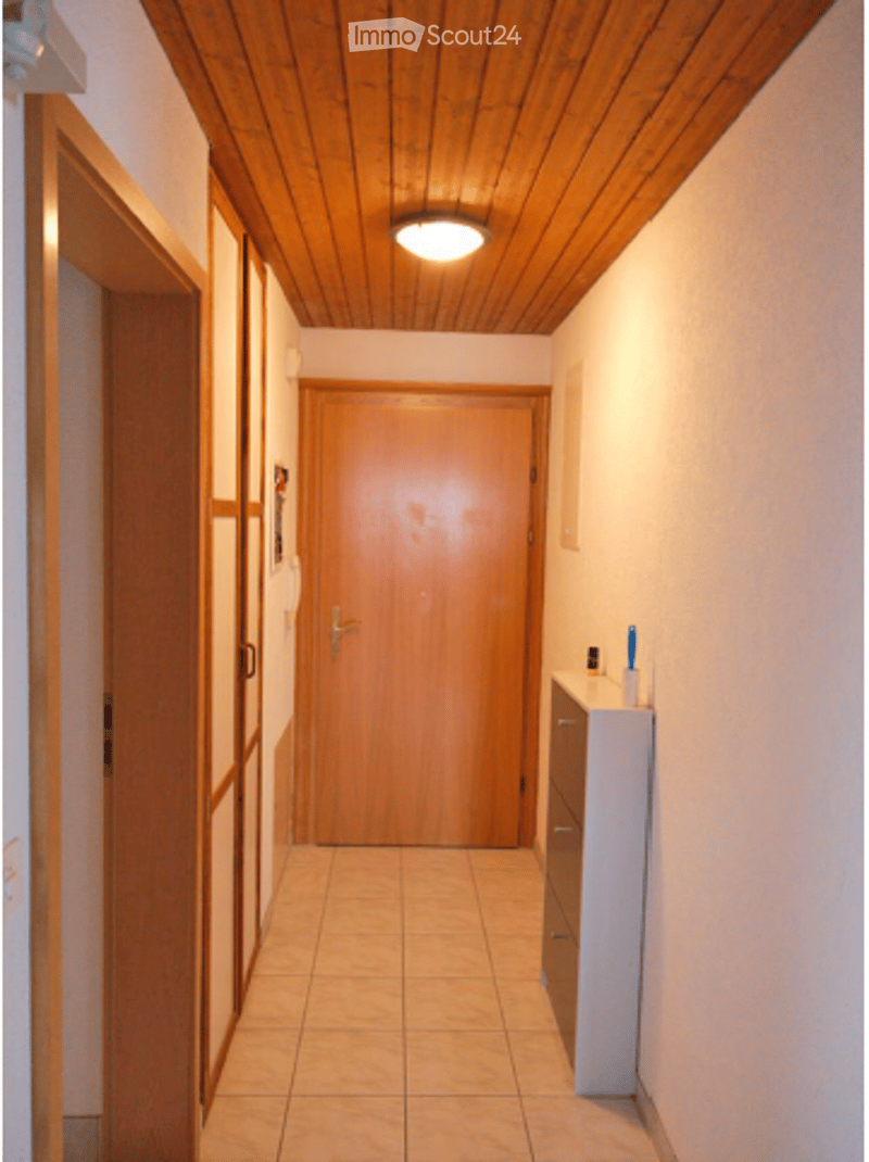 A narrow hallway with wooden ceiling, tiled floor, and white walls. Features a closed wooden door at the end, a small white cabinet with drawers on the right side, and a few small items on top of the cabinet.