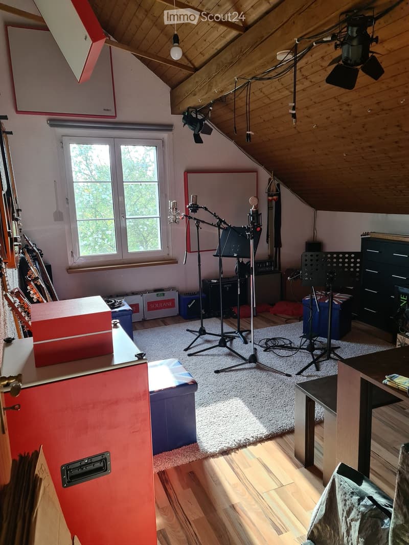 A well-lit attic room with wooden beams and slanted ceilings, featuring a microphone stand, guitar, speaker, and various musical equipment.