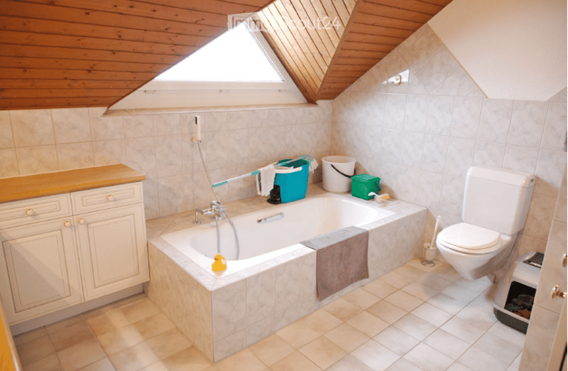 Bathroom with a bathtub, toilet, wooden vanity, tiled walls and floor, and cleaning supplies on the edge of the tub.