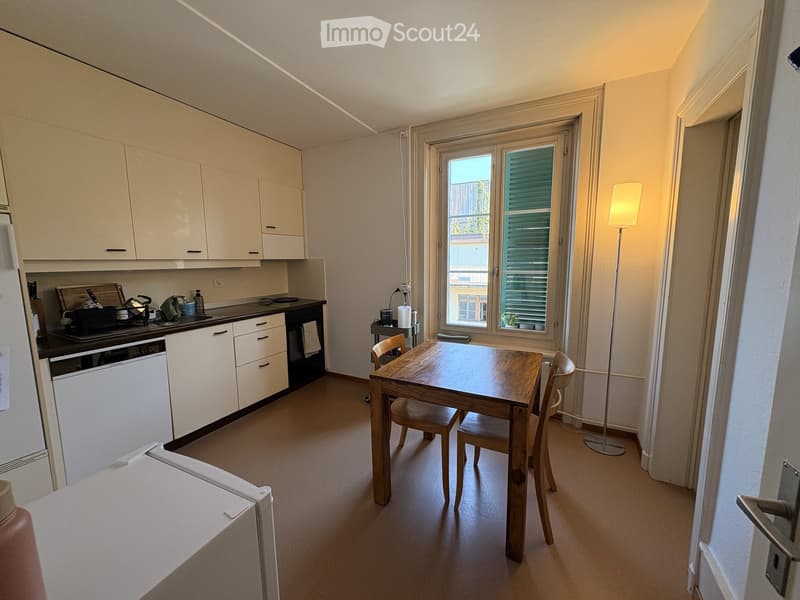 Modern kitchen with white cabinets, a wooden dining table with chairs, a refrigerator, a dishwasher, and a stove. The room features a large window with blinds and a floor lamp near the window.