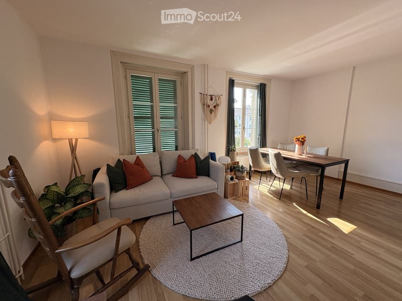 Bright living room with a grey sofa, wooden coffee table, rocking chair, dining table with chairs, and decorative plants.