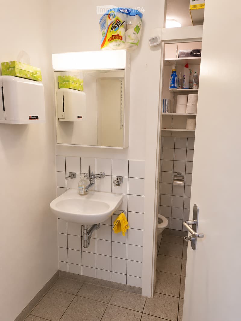 Clean bathroom with white tiled walls, a sink with a mirror above it, a toilet, shelves with cleaning supplies, and a hand dryer on the wall.