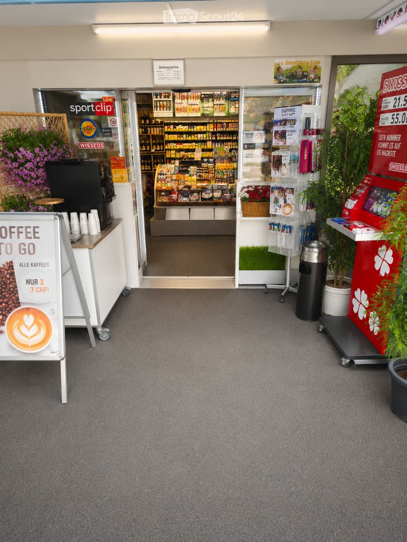 A modern, well-lit interior space with a coffee machine, a table with cups, a sign advertising coffee, and potted plants. The area is carpeted and features a view into a store with shelves of products.