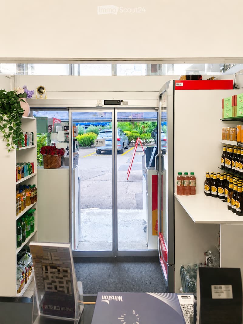 Store interior with shelves stocked with beverages, a refrigerator, a potted plant, a clock, and a brochure on the counter.