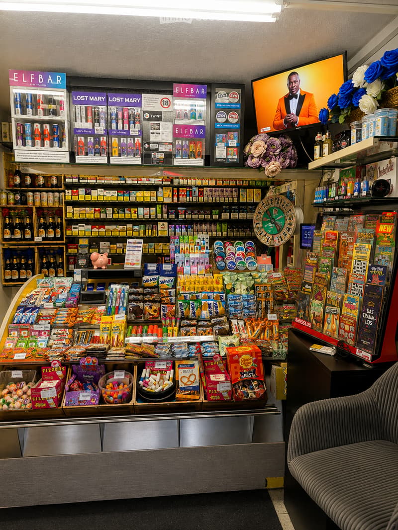 Convenience store interior with shelves stocked with various products, a television displaying a show, a sofa, and a flower arrangement on display.