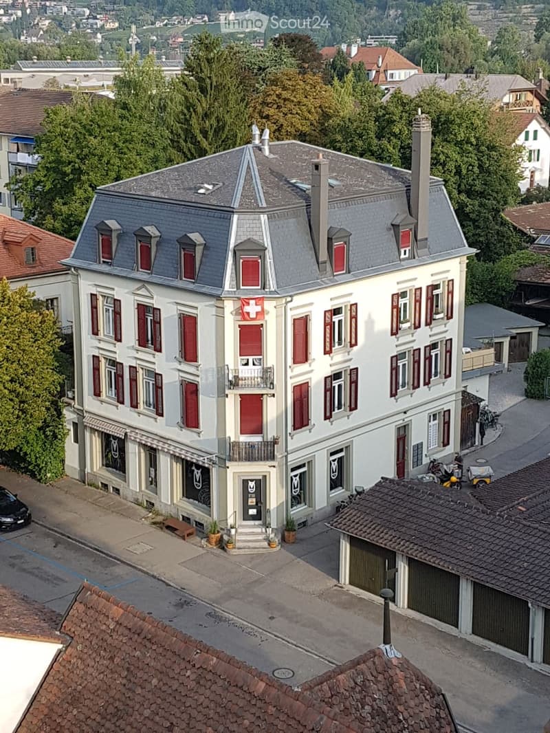 A multi-story building with a grey roof, red shutters, and a Swiss flag, surrounded by greenery and other structures.