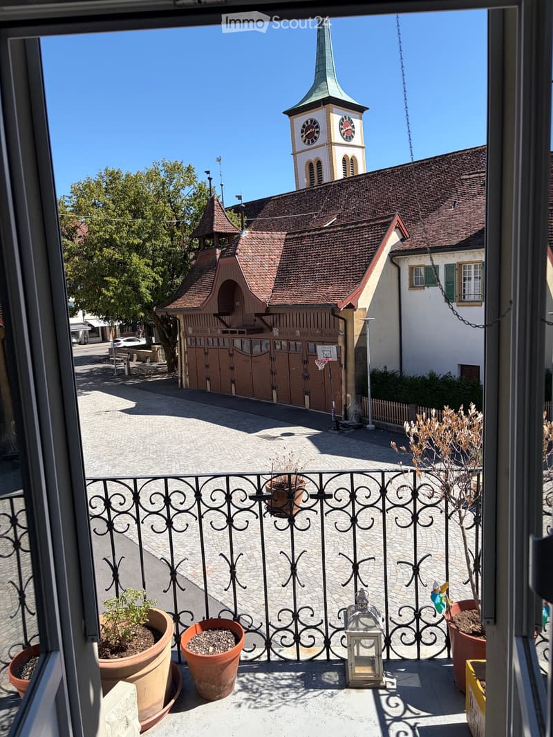 Balcony with ornate metal railing, several potted plants, and a decorative lantern. The view shows a town square with a church tower and a wooden building.