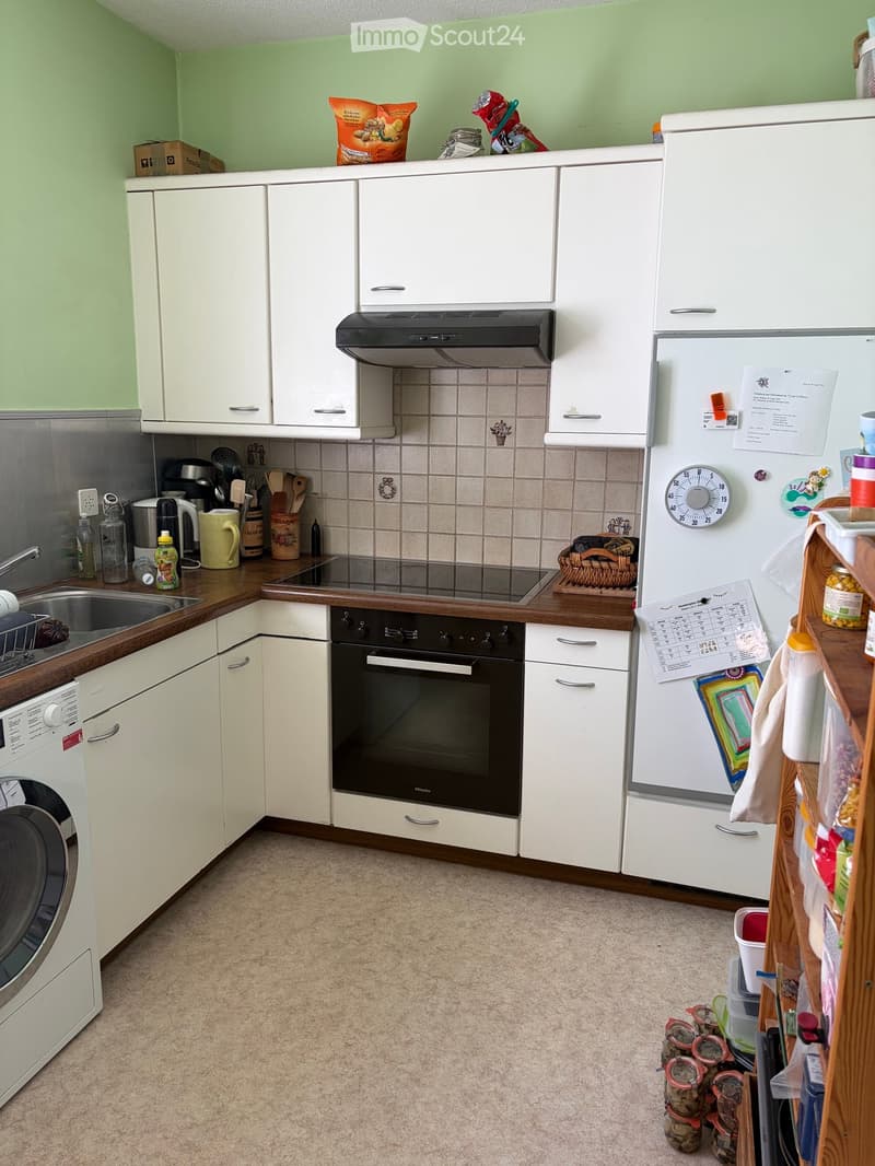 A modern kitchen with white cabinets, a stainless steel sink, a dishwasher, an oven, and a refrigerator. The countertops are wooden, and the backsplash is tiled. There are various kitchen items such as cups, bottles, and a basket on the counters.