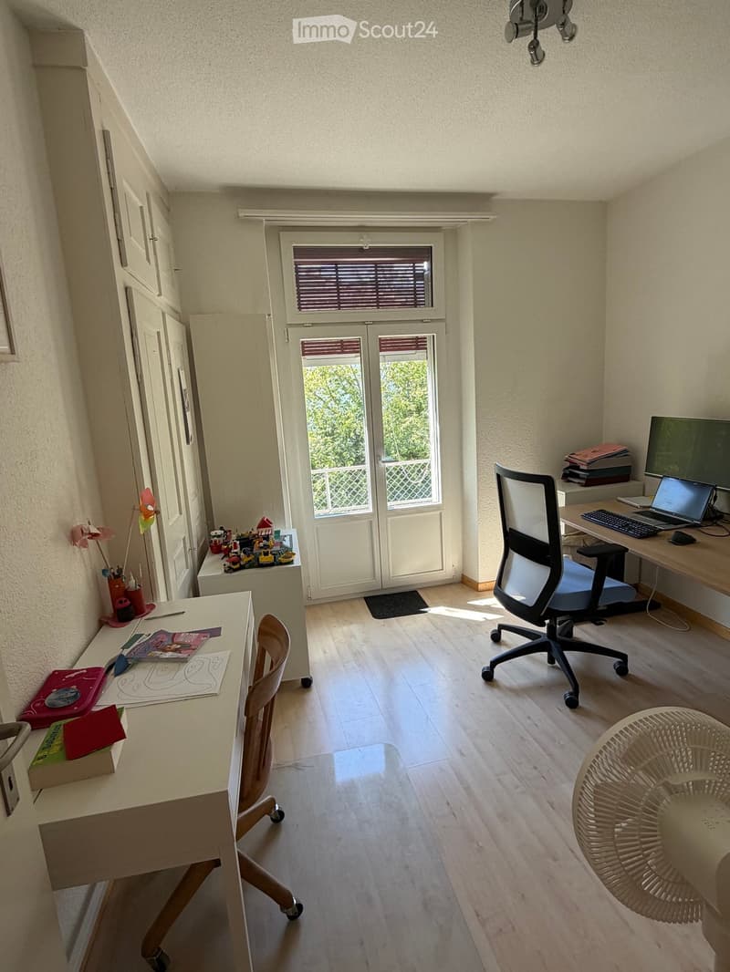 A well-lit room with wooden flooring, two desks, a chair, a laptop, a fan, and various items including books and toys.