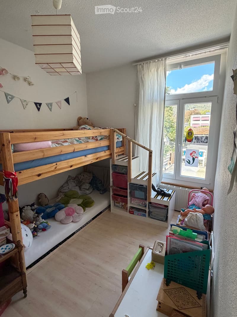 A cozy children's bedroom with a wooden bunk bed, colorful toys, a small table, and a storage unit. The room features a large window with white curtains and a view of the outdoors.
