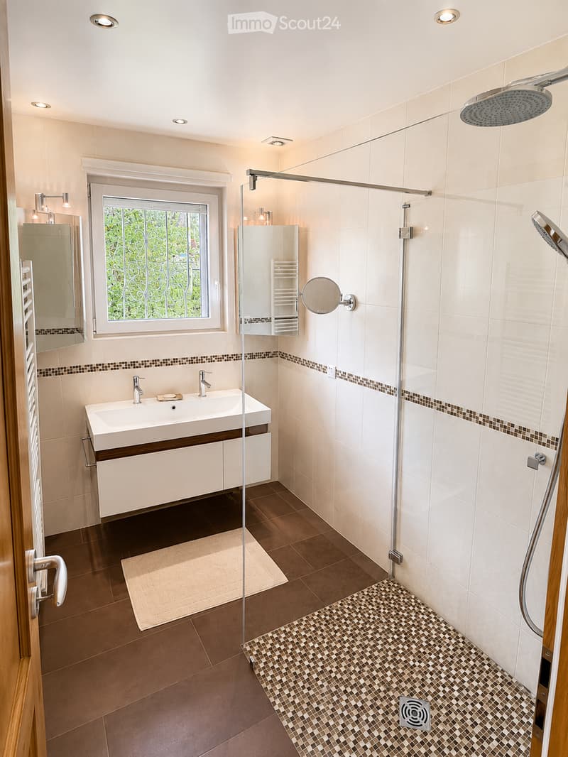 Modern bathroom with a large window, double sink, shower area, and built-in storage. Features include a glass shower enclosure, a large mirror, and a tiled floor.