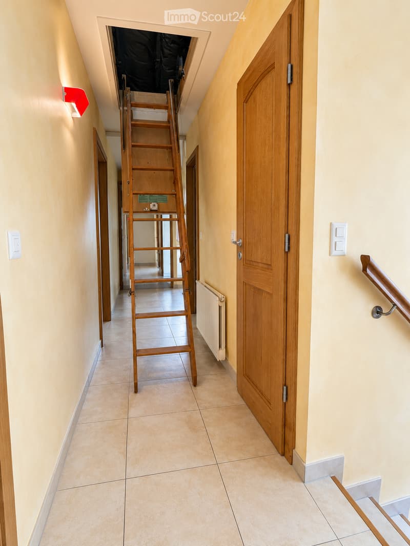 A narrow hallway with beige tiled flooring, light yellow walls, and wooden doors. A wooden ladder is positioned against the wall, leading to an attic space. A handrail is visible on the right side.