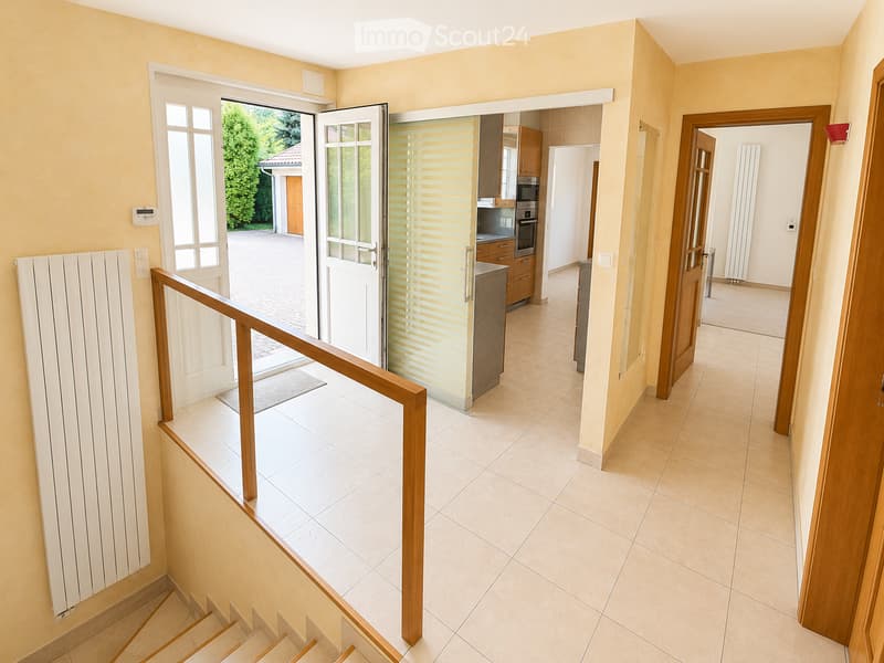 Spacious hallway with tiled flooring, wooden staircase, and open doors leading to a kitchen and outdoor area.