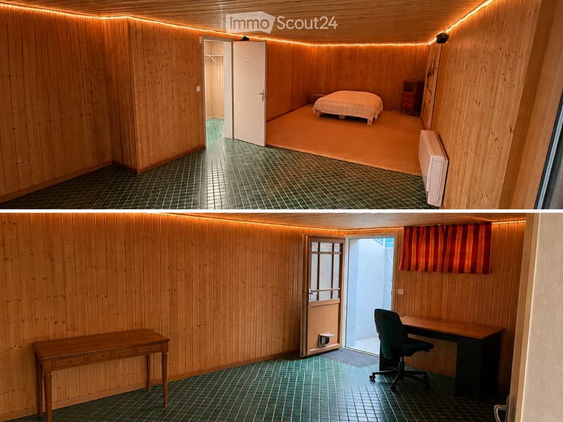 Two connected rooms with wooden walls and green tiled flooring. The first room contains a bed, nightstand, and a small table. The second room features a desk, chair, and a curtained window.
