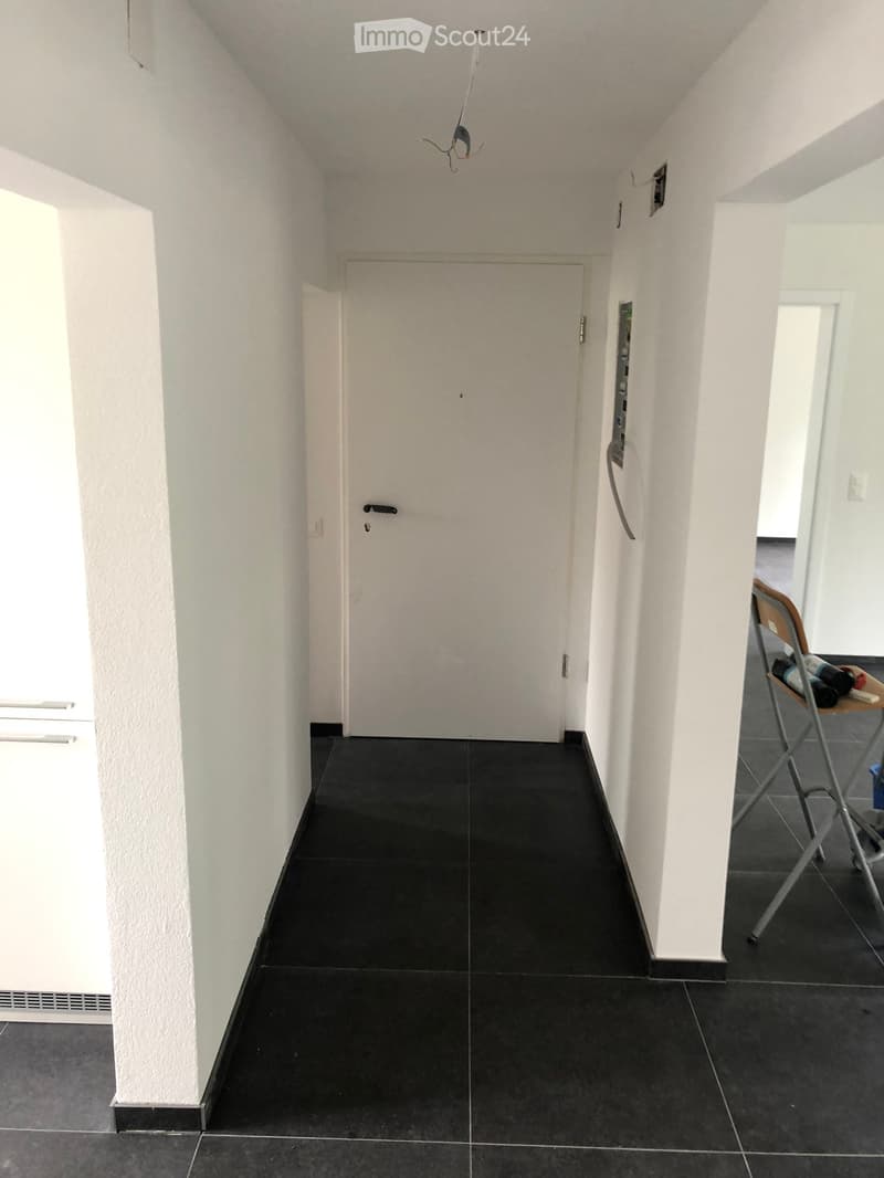 Empty hallway with white walls, black tiled floor, and a closed door at the end. A small folding chair with items on it is visible to the right.