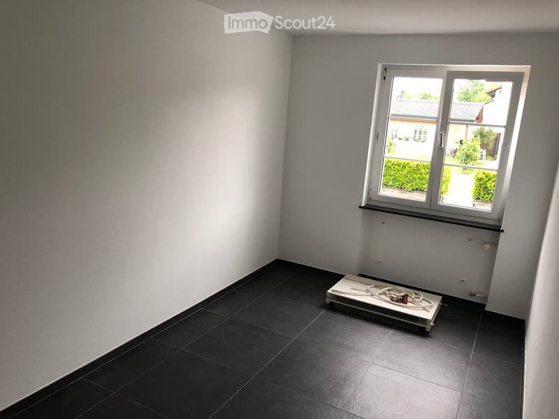 Empty room with dark tiled floor, white walls, and a window showing a view of a garden and neighboring house.