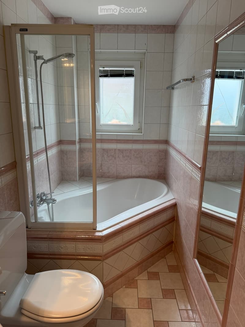 Modern bathroom with a bathtub, toilet, shower, and large windows providing natural light. The walls are tiled with a decorative border, and the floor is tiled in a warm, neutral tone.