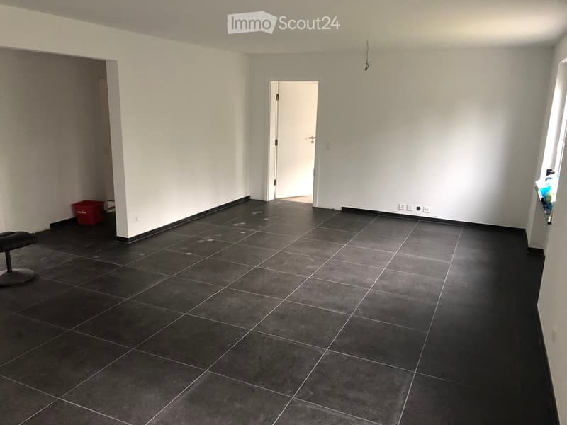 Empty room with dark tiled floor, white walls, and a single door leading to another space. A small black stool and a red storage bin are visible.