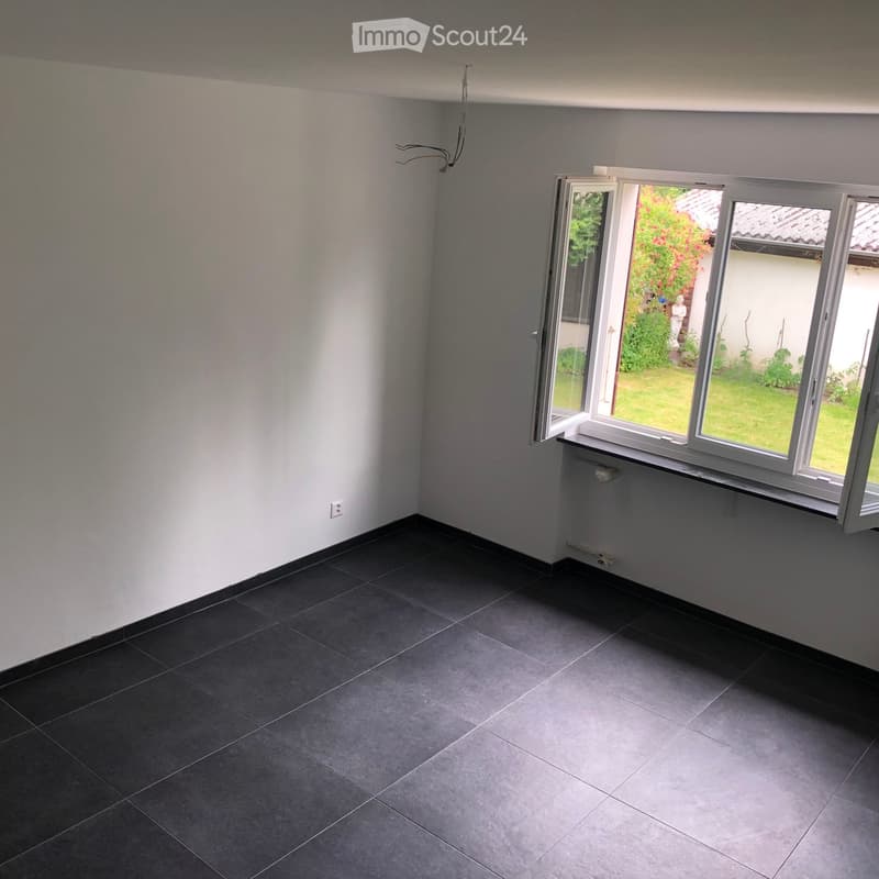 Empty room with dark tiled floor, white walls, and large windows opening to a garden view.