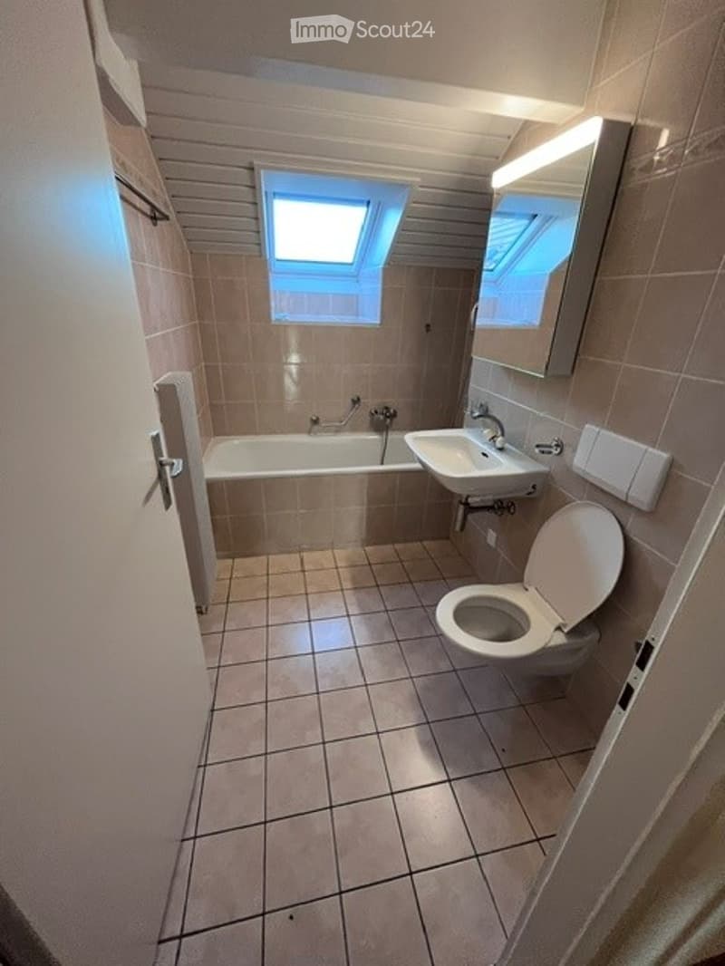 Bathroom with tiled walls and floor, bathtub, sink, toilet, and a mirror above the sink. A small window is visible above the bathtub.