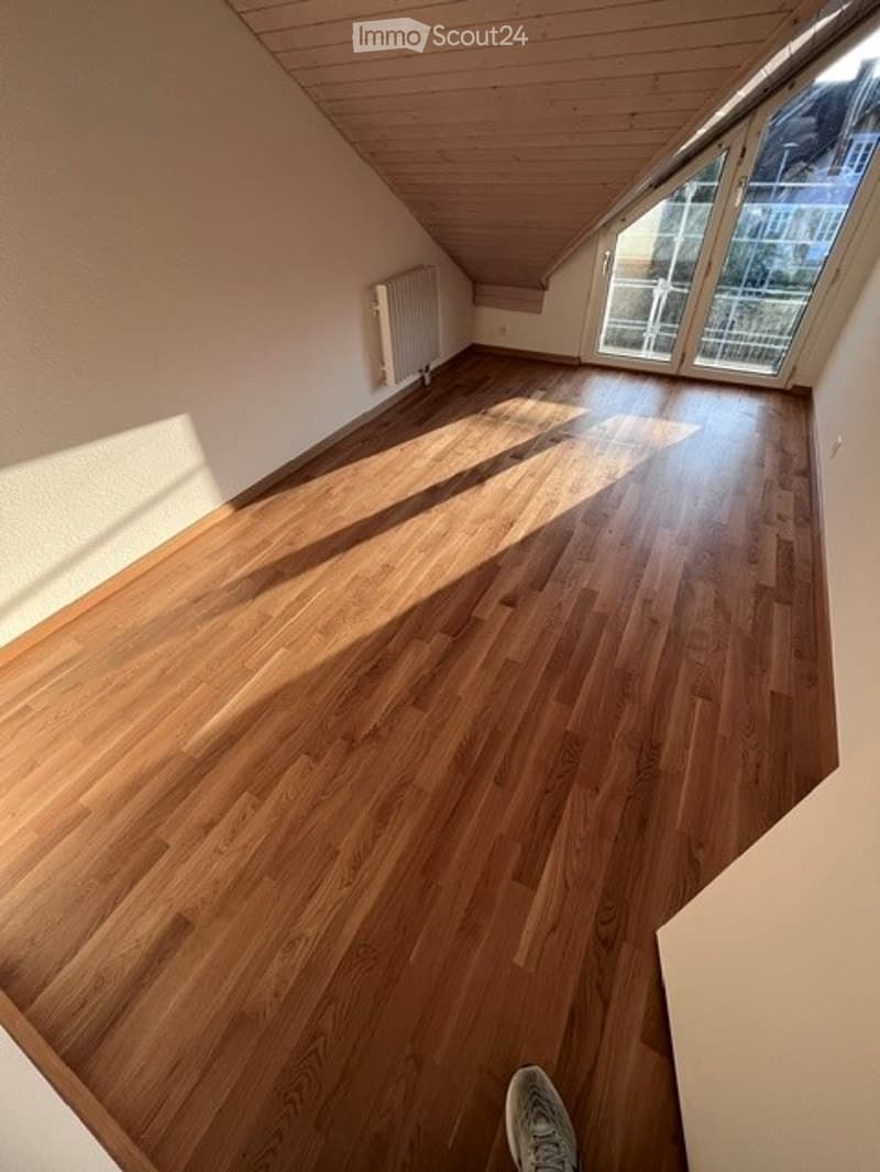 Empty attic room with wooden flooring, slanted wooden ceiling, large window offering a view, and a radiator on the wall.