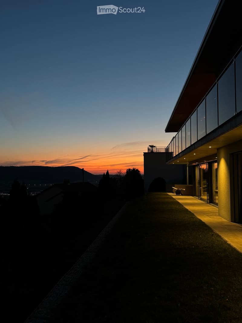 Modern two-story house with large windows and a balcony, illuminated at dusk with a view of the sunset and surrounding landscape.