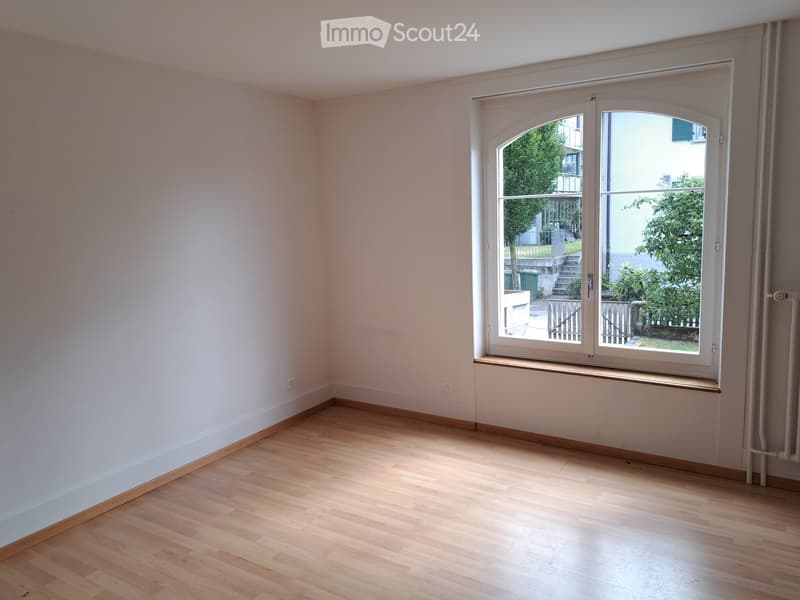 Empty room with light wooden flooring, white walls, and a large window with a view of a garden area.