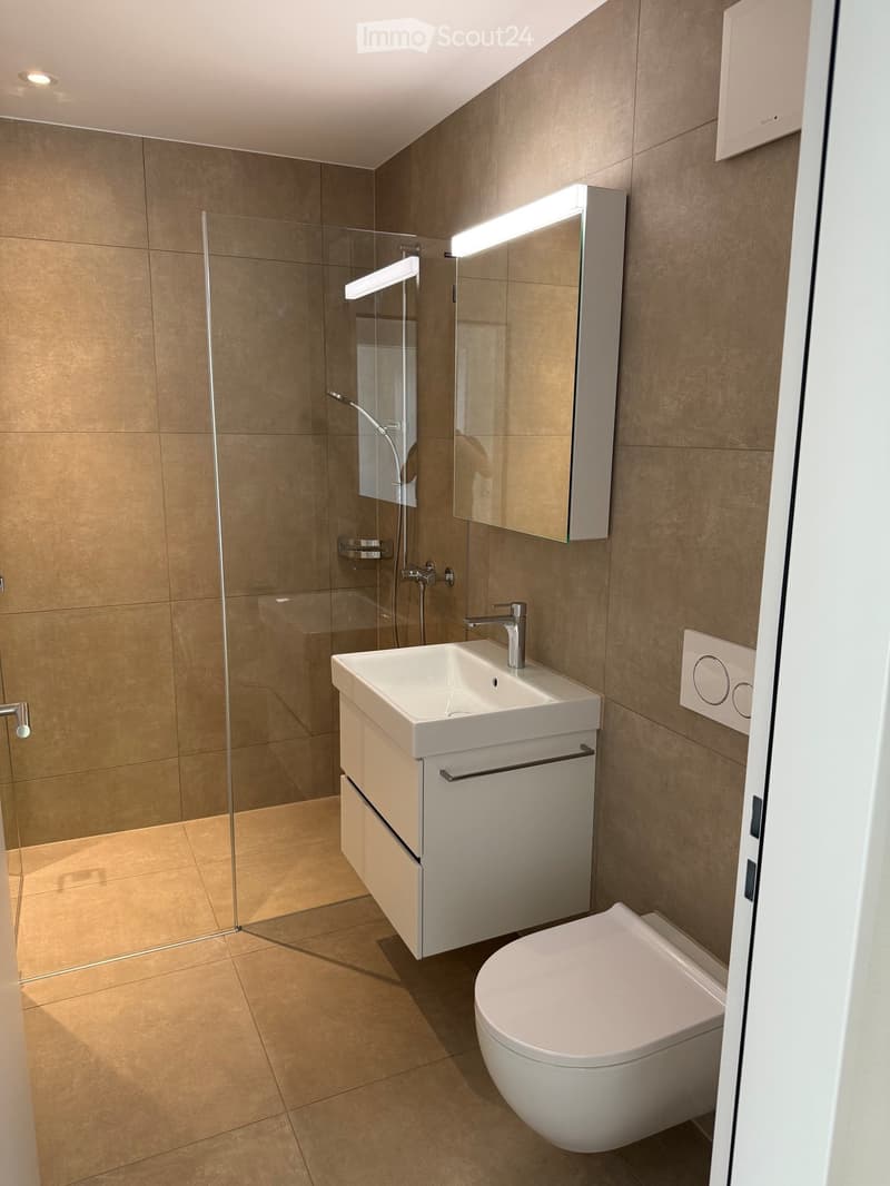Modern bathroom with beige tiles, white sink, toilet, and glass-enclosed shower with a modern faucet and mirror.