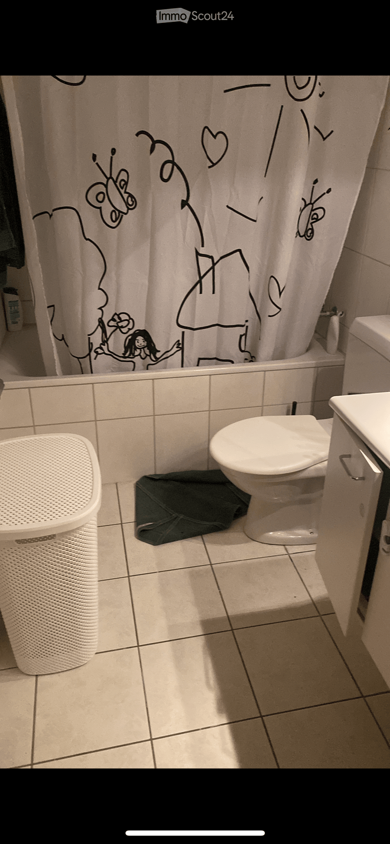 Bathroom with a toilet, sink, shower curtain featuring cartoon characters, and a white trash can. The floor is tiled, and a dark-colored bag is placed near the toilet.