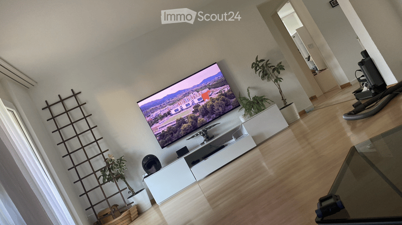 Modern living room with a large flat-screen TV mounted on the wall, a sleek white TV stand, a glass coffee table, and several potted plants. The room features light wooden flooring and white walls.