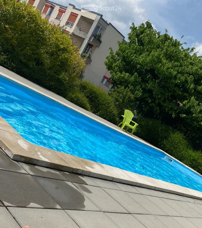 A rooftop swimming pool with clear blue water, surrounded by greenery and a small green plastic chair.
