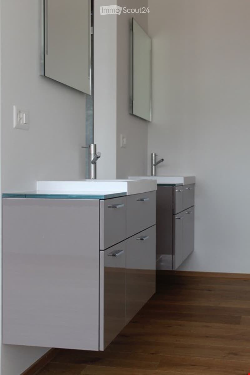 Modern bathroom with two sinks, sleek grey cabinets, glass countertop, and mirrors above the sinks.