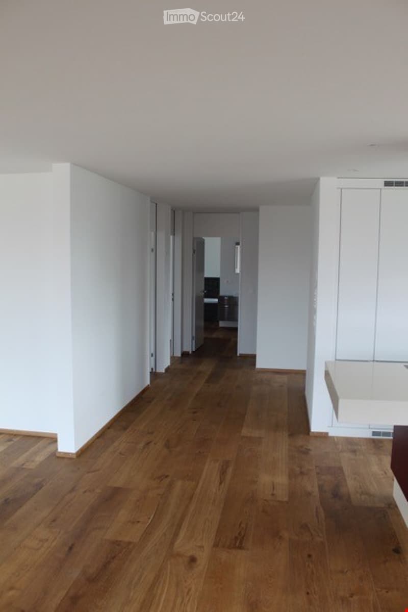 Empty hallway with light wooden flooring and white walls, leading to a partially visible room with a dark-colored cabinet.