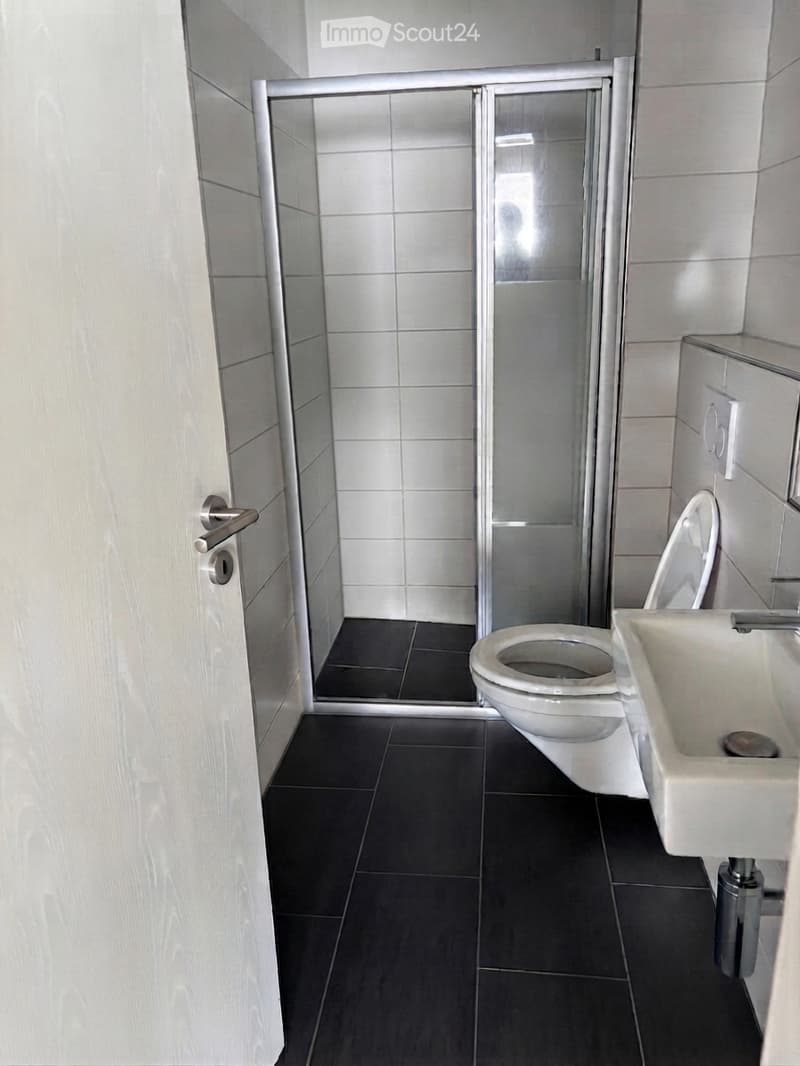 Modern bathroom with white tiles, black floor tiles, a toilet, a sink, and a glass-enclosed shower area.