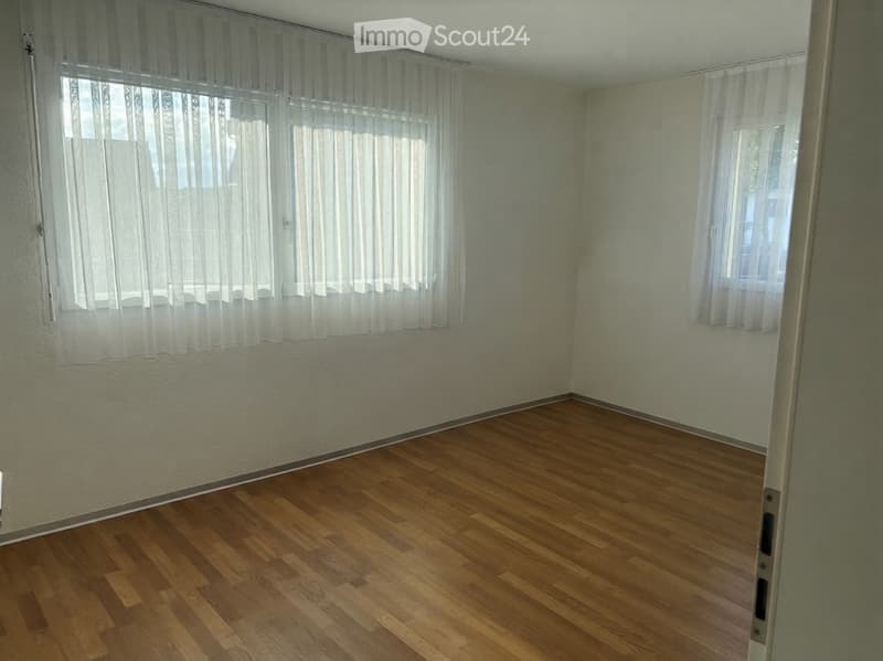 Empty room with wooden flooring and white walls, featuring two windows with sheer curtains.