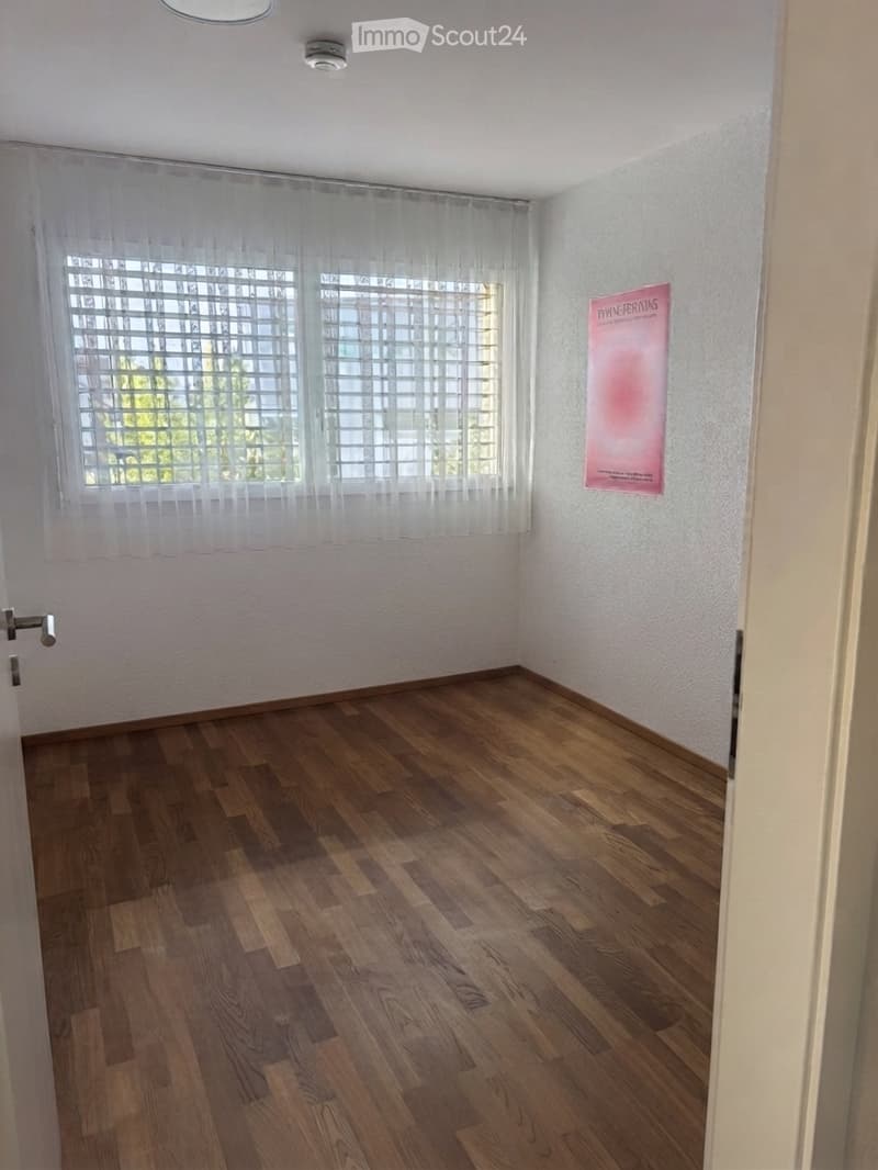 Empty room with wooden flooring, white walls, large windows with blinds, and a single poster on the wall.