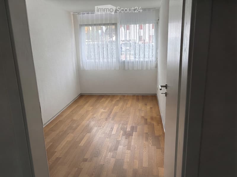 Empty room with wooden flooring and white walls, featuring a window with sheer curtains and a door.
