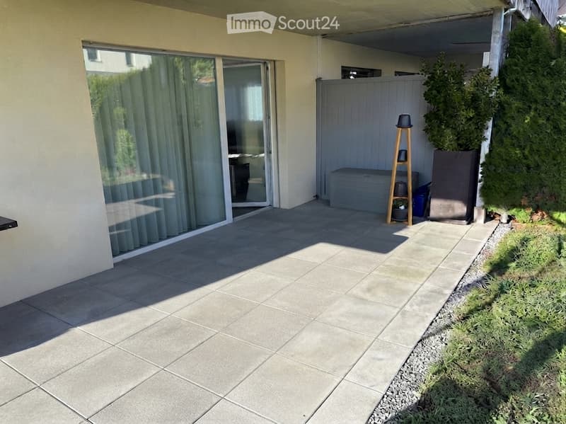 A modern patio area with large sliding glass doors leading to a room, tiled floor, and a small garden with shrubs and a tree in a planter.