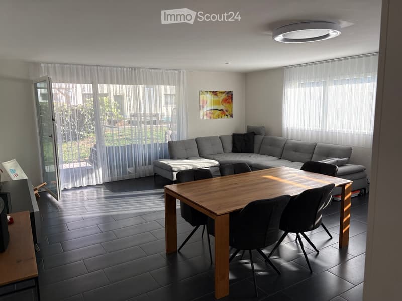 Modern living room with grey sectional sofa, wooden dining table, black chairs, large windows with sheer curtains, and a round ceiling light.