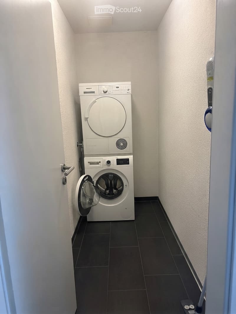 Compact bathroom with a stacked washing machine and dryer unit, dark tiled floor, white walls, and a door with a silver handle.