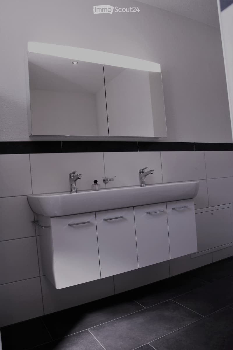 Modern bathroom with a white sink, two chrome faucets, a mirrored cabinet with built-in lighting, and white cabinets with silver handles.