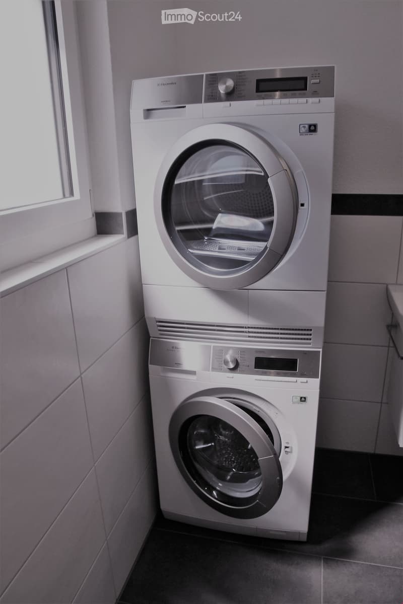 A compact bathroom with a stacked washing machine and dryer unit, featuring a control panel and a window for natural light.
