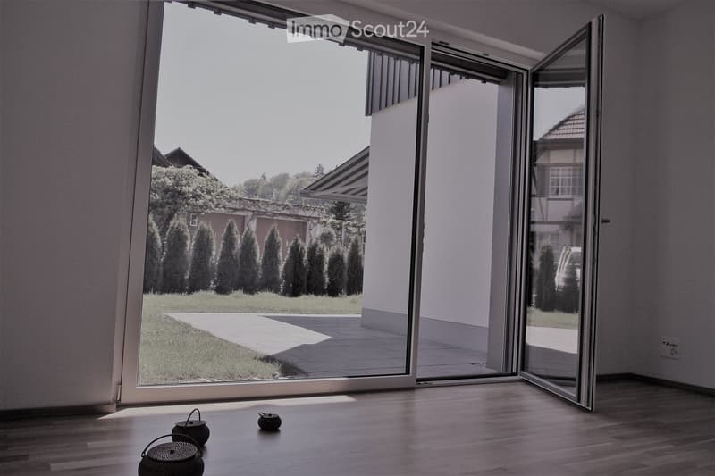 Empty room with wooden flooring and large sliding glass doors opening to a garden with neatly trimmed trees and a paved pathway.